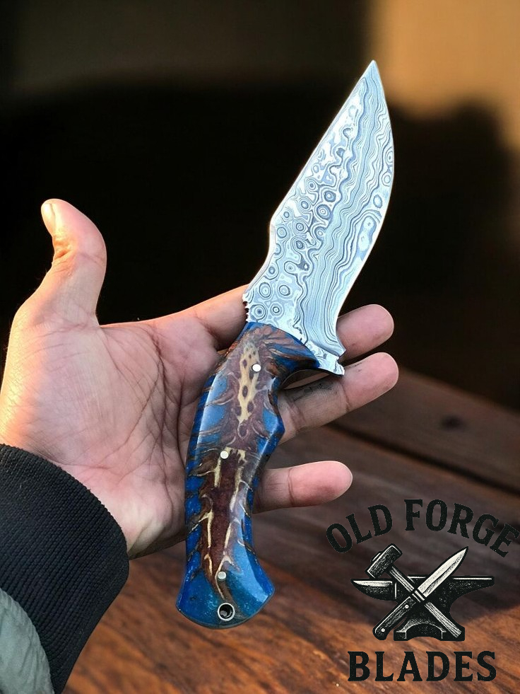 Hand holding a knife with a decorative blade and blue handle, branded 'Old Forge Blades'.